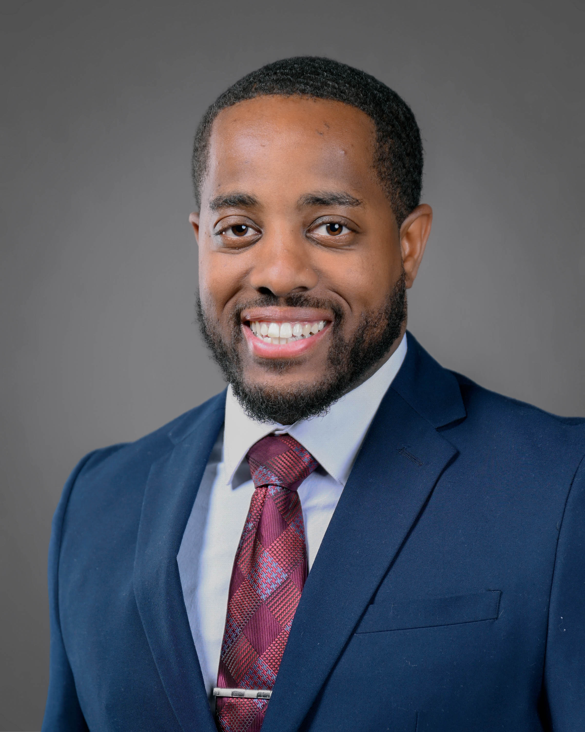 Man smiling with short curly brown hair and a short beard wearing a blue suit with a red tie